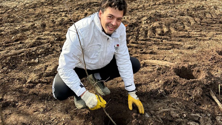 Man knielt bij het planten van een jonge boom in het veld. Draagt jas, handschoenen en donkere broek.