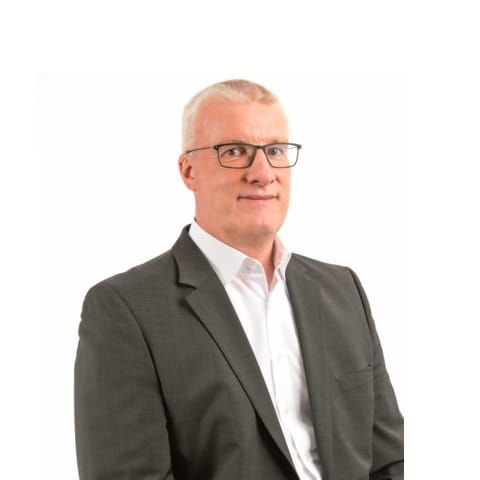 Man met kortgeschoren haar, bril en donker pak voor witte achtergrond.