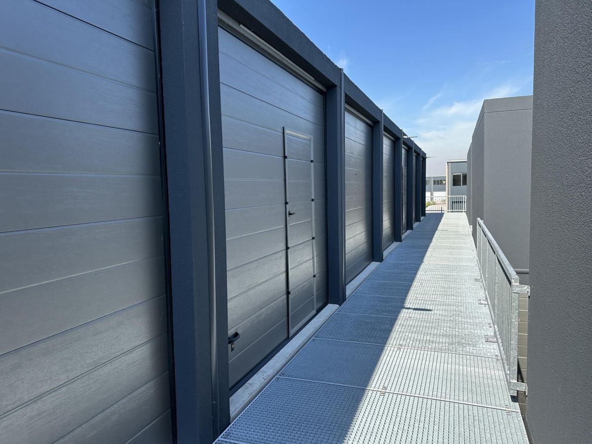 Rij grijze garages met sectionele deuren en smalle metalen brug ervoor onder blauwe lucht.