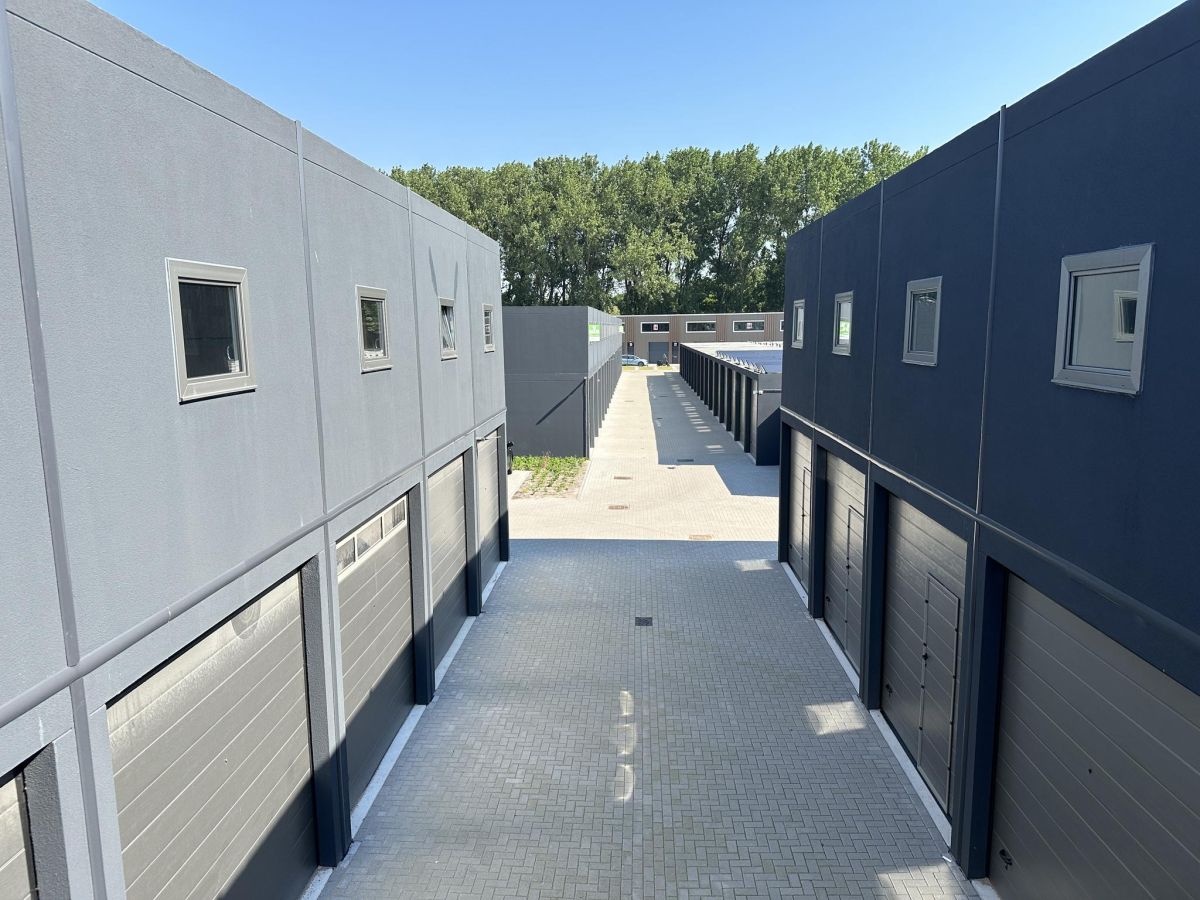 Rij grijze garages met roldeuren aan een geplaveide rijbaan. Op de achtergrond een gebouw en bomen.