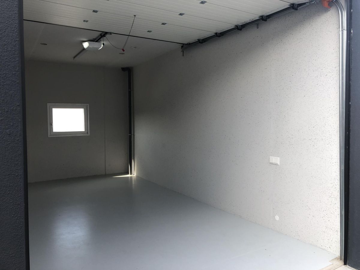 Lege ruimte met lichtgrijze muren en vloer, raam links, garage rechts, verlichting aan het plafond.