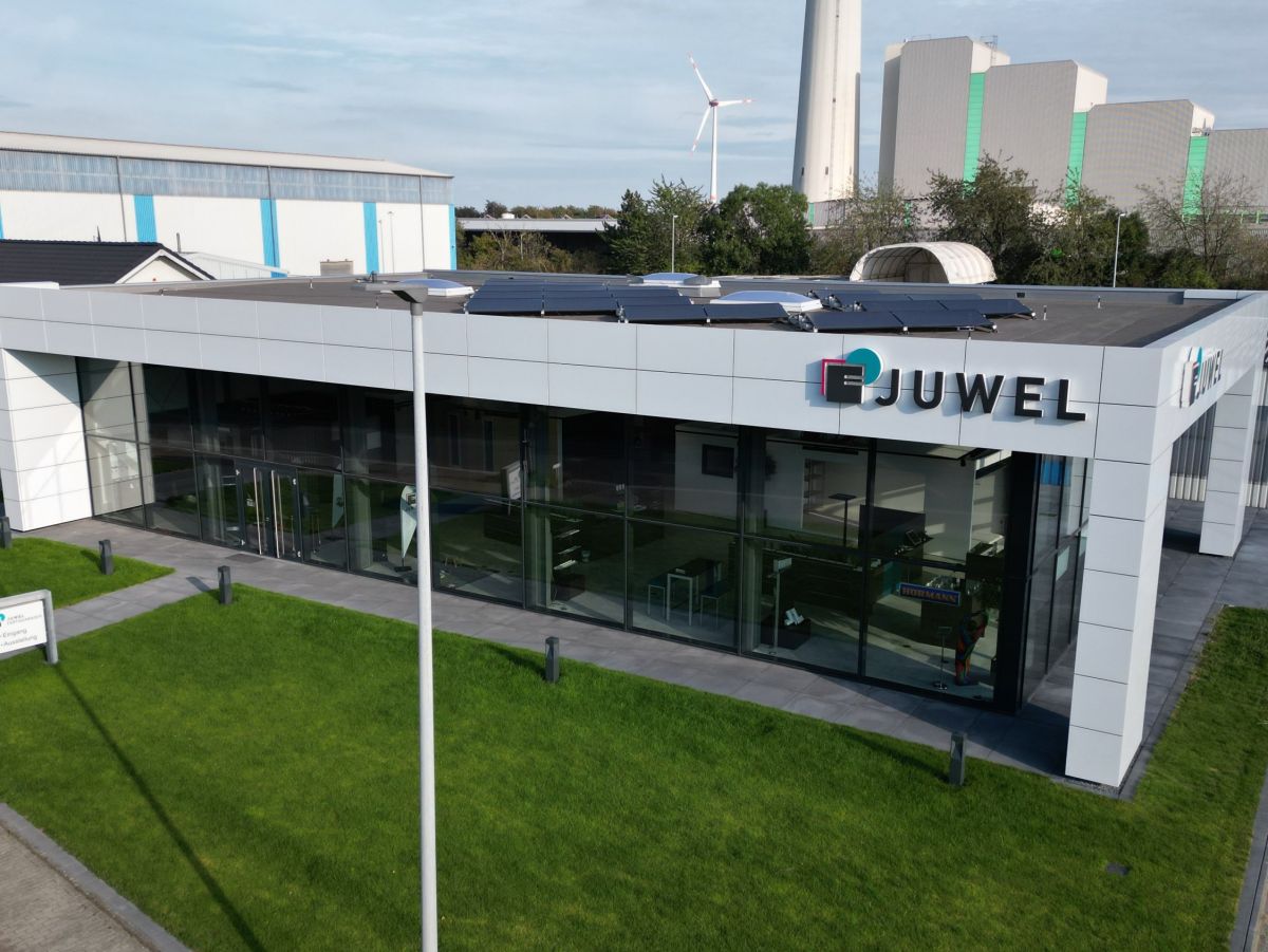 Laag gebouw met glazen gevel en de tekst 'JUWEL'. Windturbine en industriële installaties op de achtergrond.