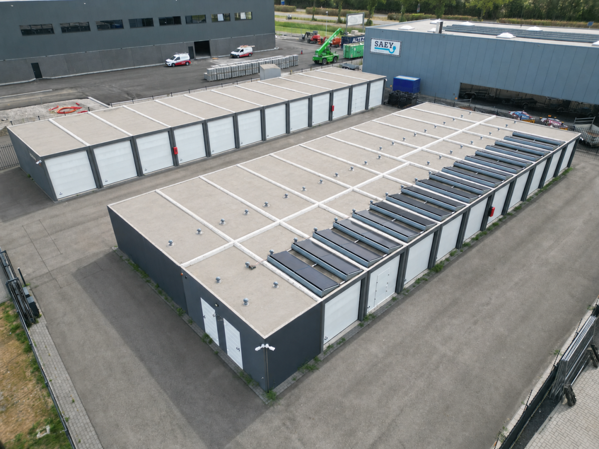 Luchtopname van een magazijn met plat dak, zonnepanelen en een geplaveide voorplaats. Op de achtergrond andere industriële gebouwen.