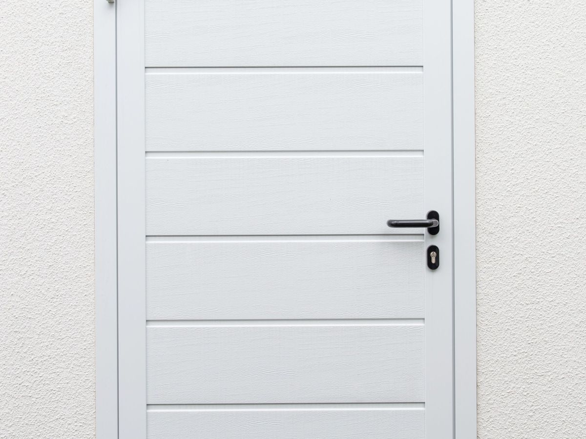 Witte deur in een lichte muur. Deur met horizontale groeven en zwarte handgreep. Onder grind en schaarse vegetatie.