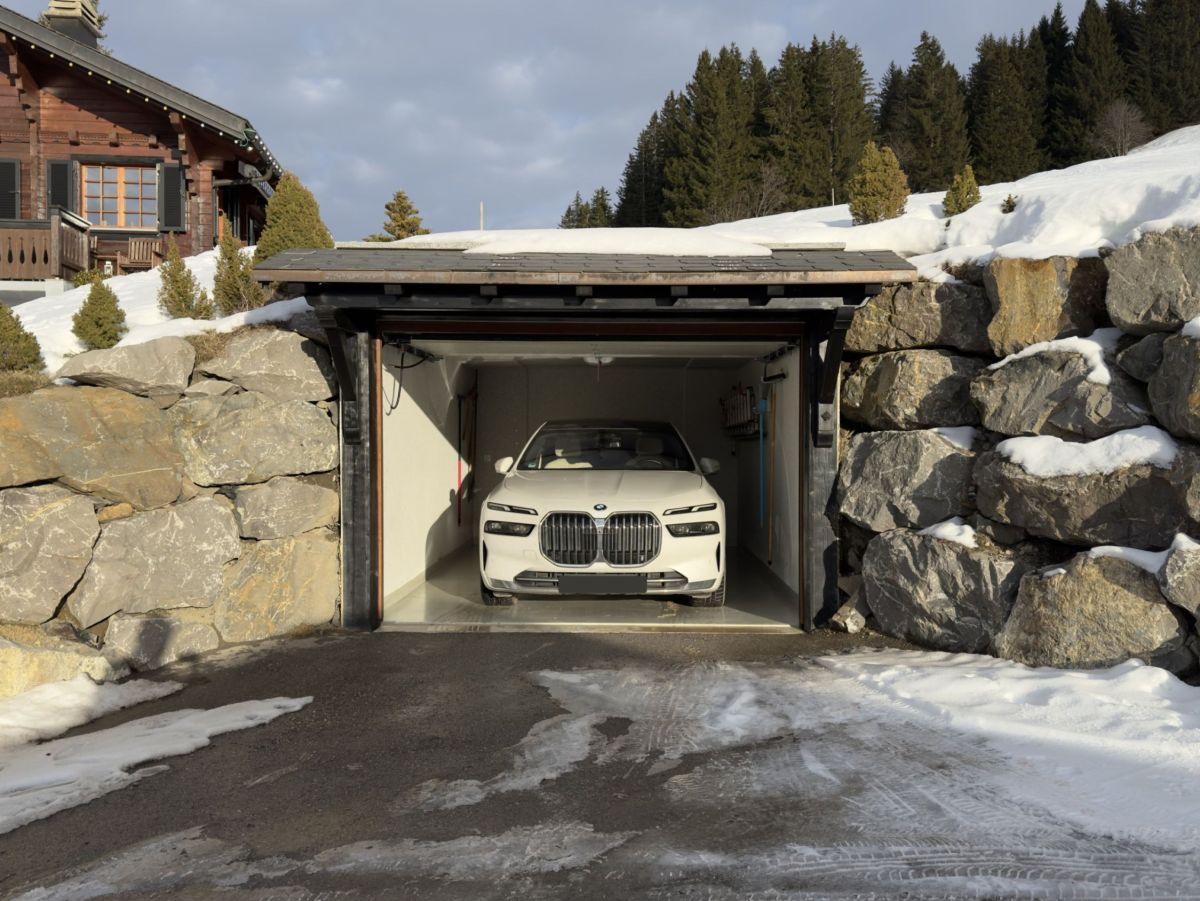 Witte BMW rijdt in een garage, ingebouwd in een rotswand. Op de achtergrond een chalet en besneeuwd landschap.
