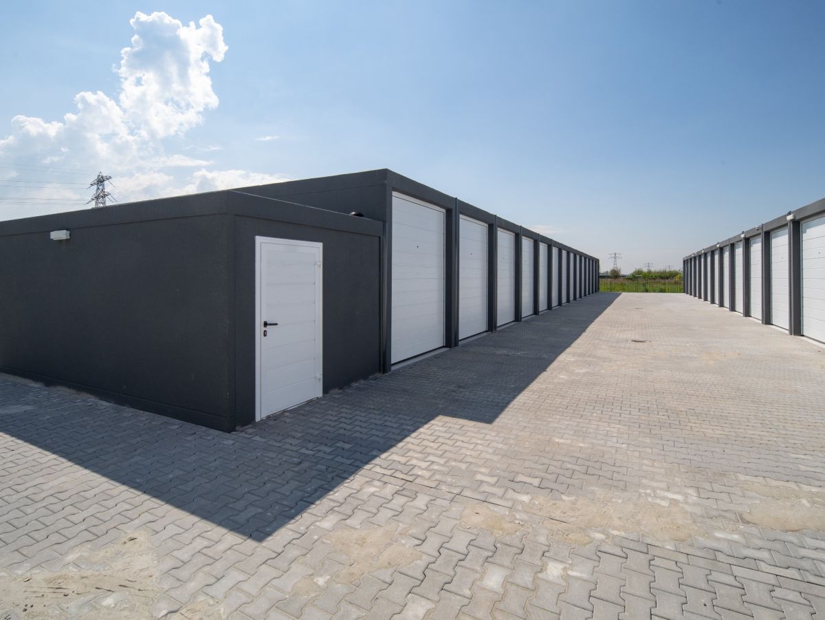 Rij grijze garages met witte deuren op een geplaveide ondergrond onder een blauwe lucht met wolken.
