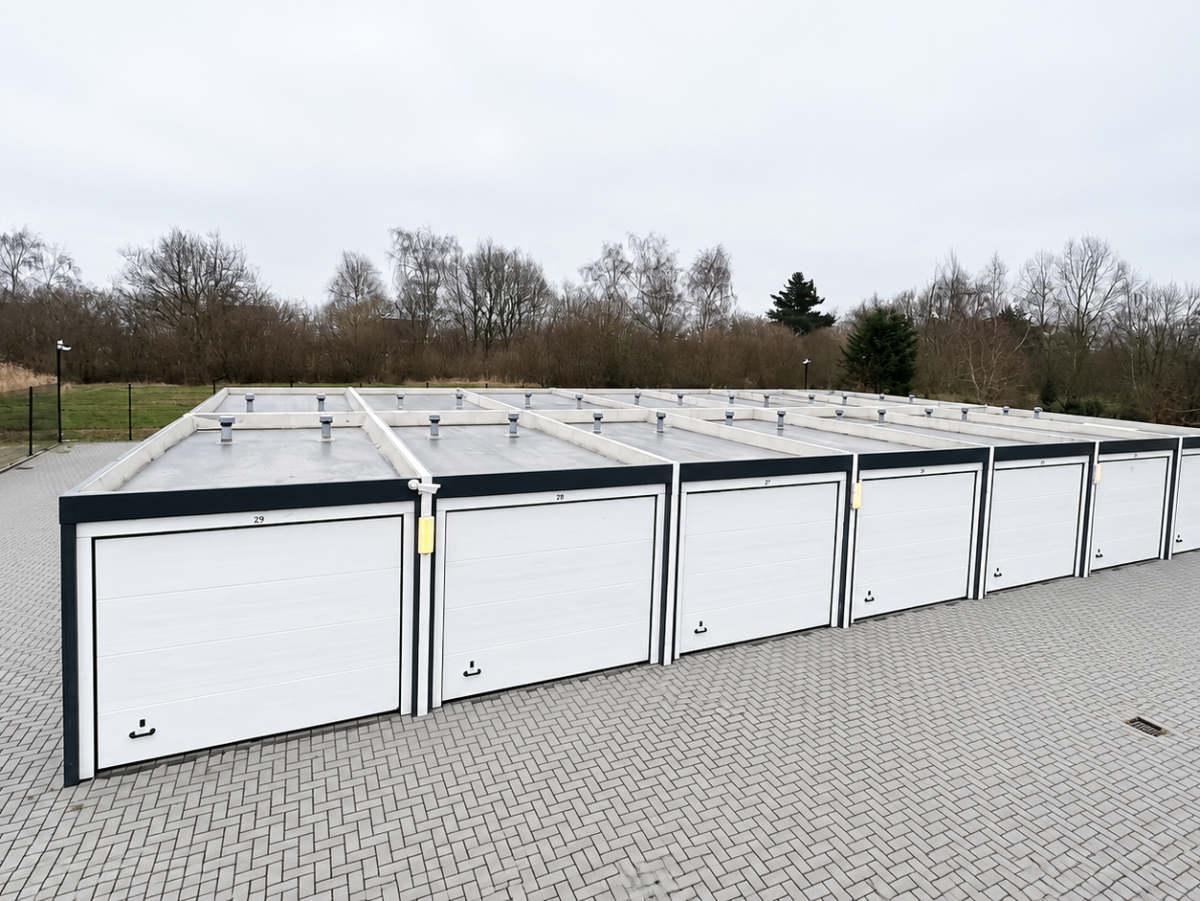 Rij garages met witte garagedeuren op een betegelde oprit. Op de achtergrond bomen en een gebouw.