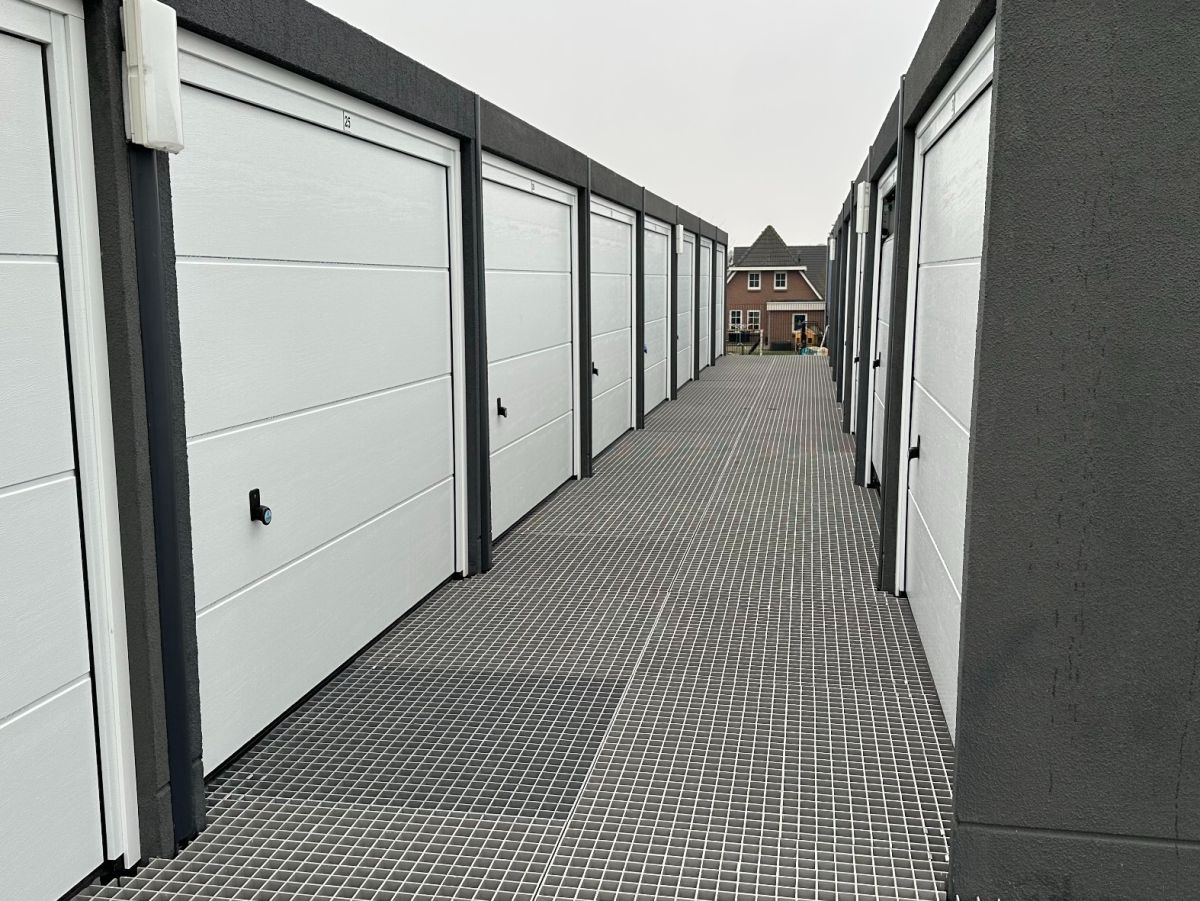 Rij grijze garages met witte deuren aan een geplaveide weg. Op de achtergrond een woonhuis.