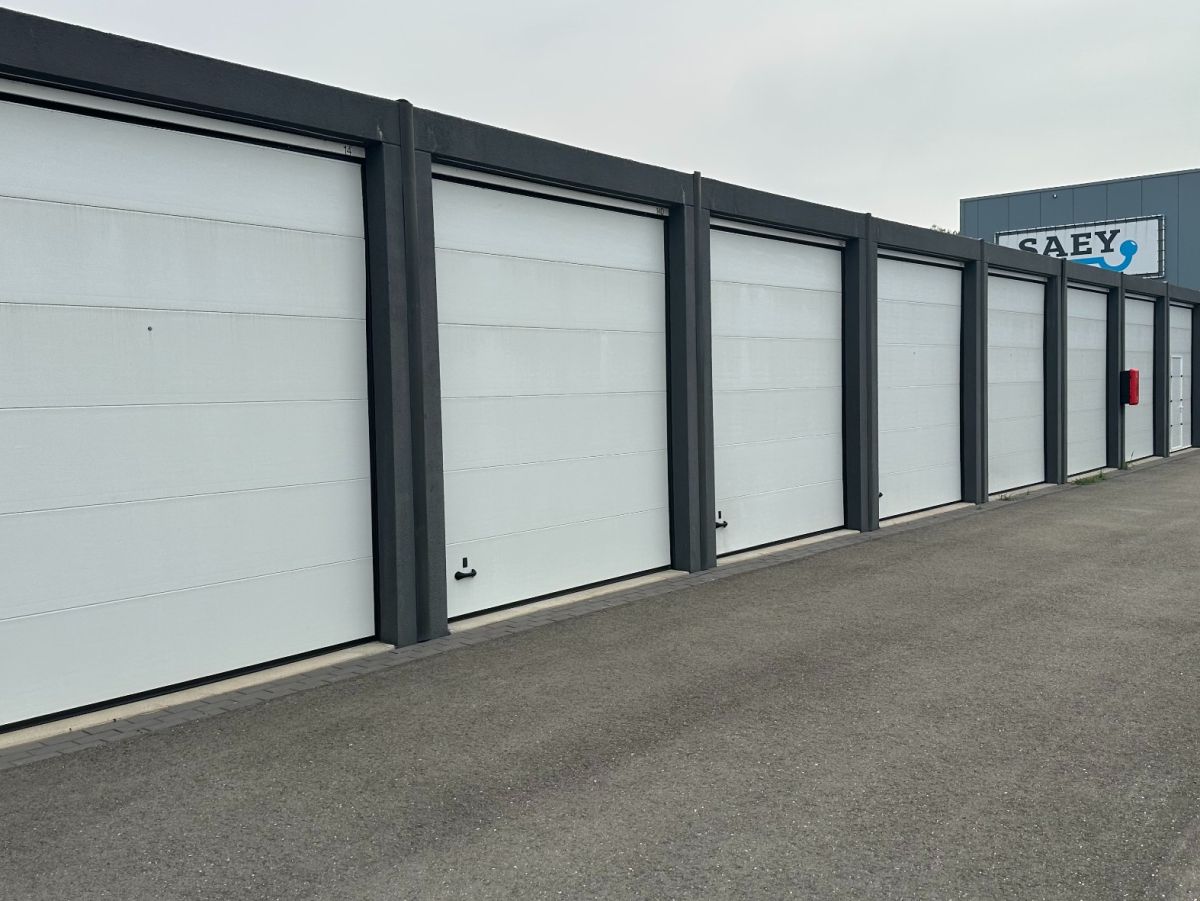 Rij witte garagedeuren met grijze frames bij een opslagloods. Op de achtergrond een blauw gebouw met logo.