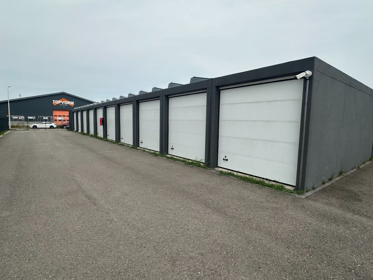Rij grijze garages met witte roldeuren op een geasfalteerde binnenplaats. Op de achtergrond een donkerder gebouw met logo.