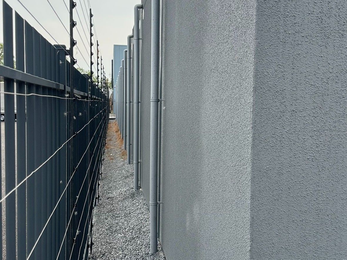 Hoge grijze betonnen muur langs een smal grindpad. Bovenop een hek met elektrische draden.