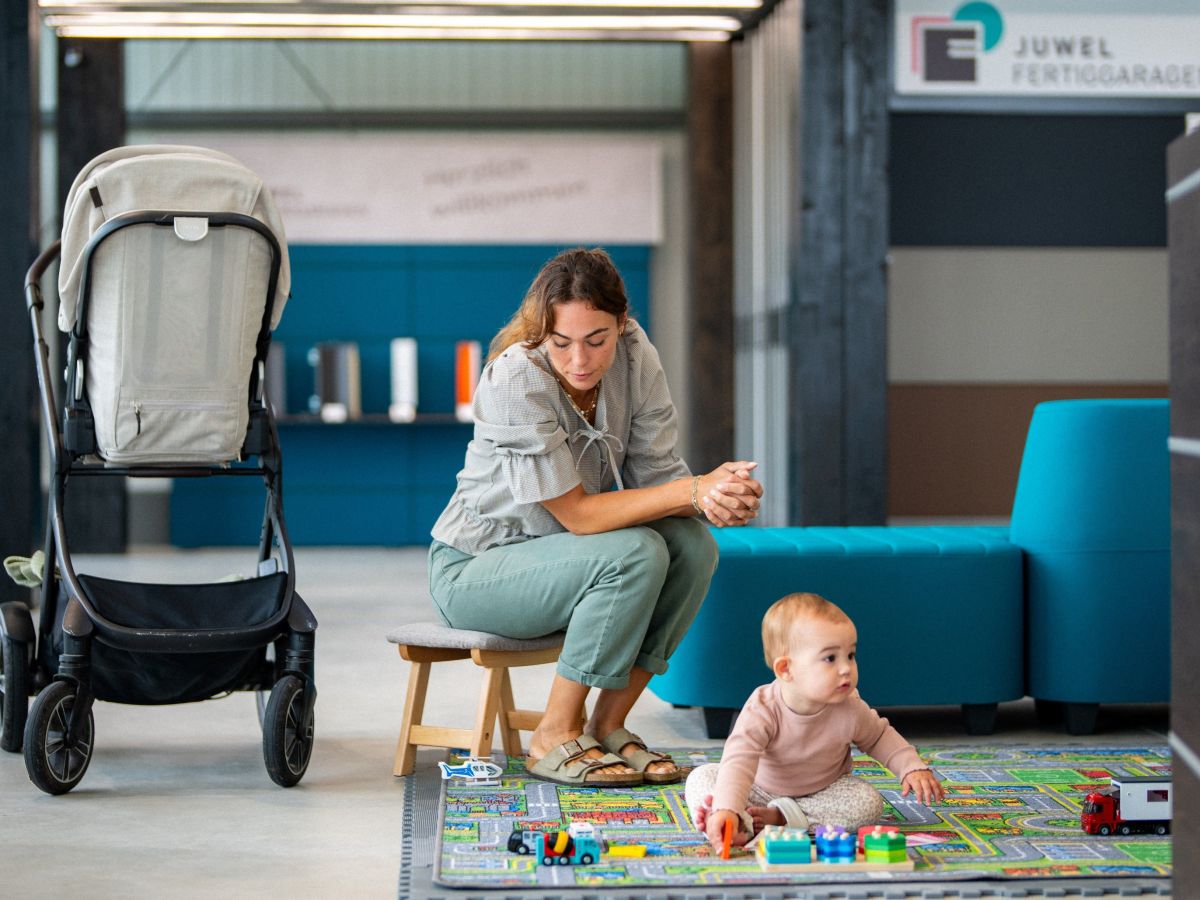 Vrouw zit voor baby op speelmat. Links kinderwagen, op de achtergrond reclamebanner.