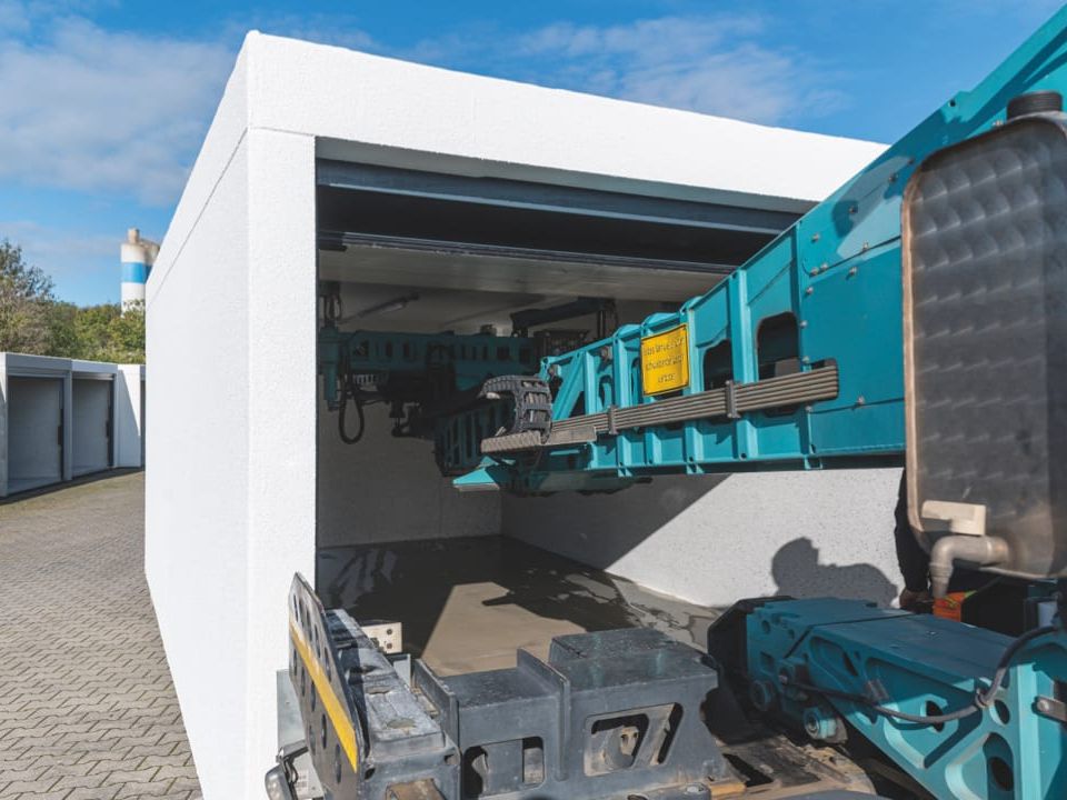 Heldergrijs garagegebouw met open deur, gedeeltelijk zichtbaar turquoise speciaal voertuig erin.