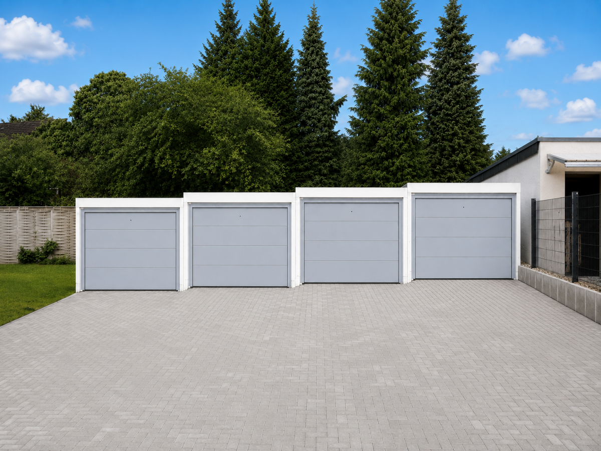 Rijenwoning met vier grijze garagedeuren en geplaveerde voortuin, gras en bomen ervoor.