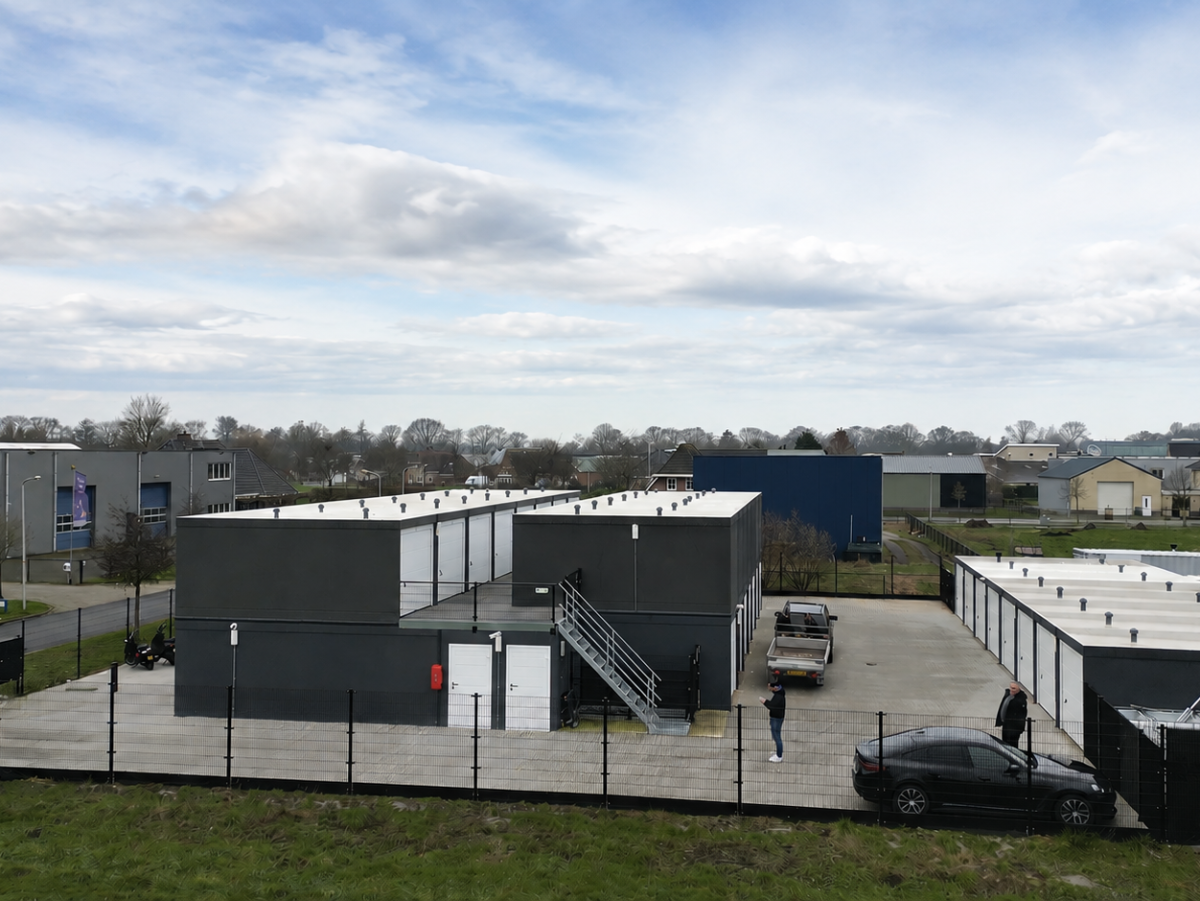 Industrieterrein met meerdere rechthoekige gebouwen, omheind door een zwart hek. Bewolkte lucht.