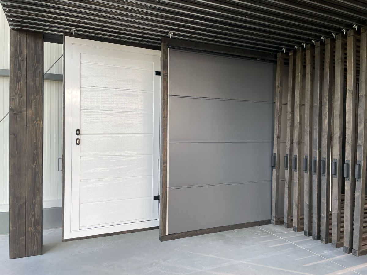 Gevel van een gebouw met witte deur links en grijze garagedeur rechts. Moderne houten constructie als overkapping.