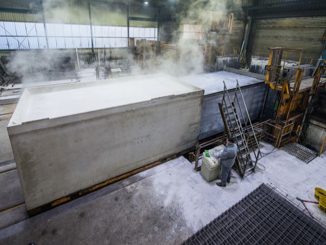 Betonfabriek: Werker naast grote, stoomende betonkuip. Op achtergrond verdere betonmallen en industrie-installaties.