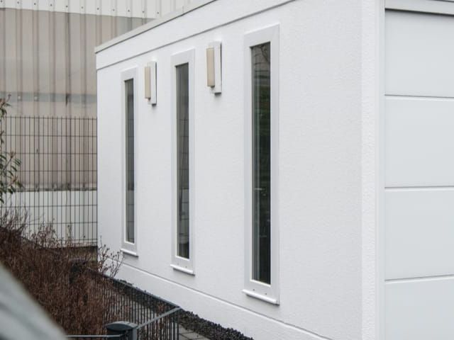 Witte, rechthoekige container met drie smalle ramen. Achtergrond: metalen hal en hek.
