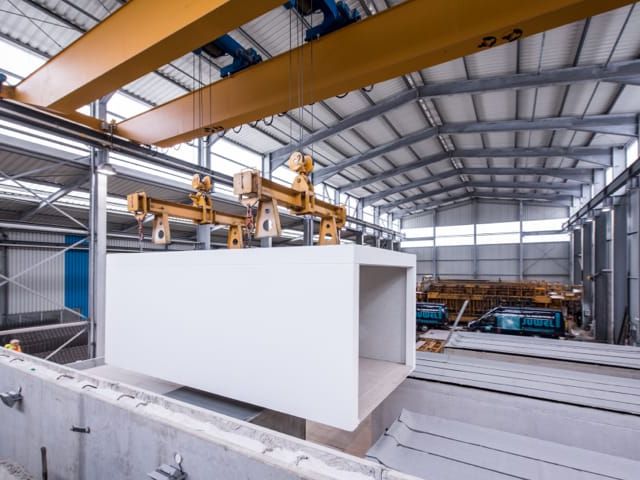 Fabriekshal. Grote betonblok wordt door kraanarmen opgetild. Op de achtergrond meer betonelementen.