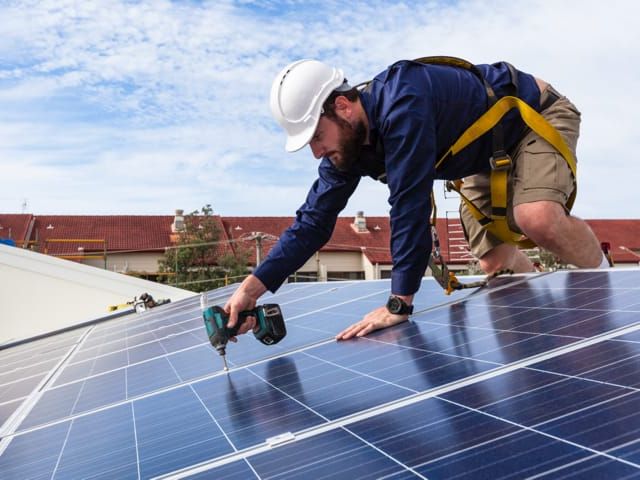 Man in veiligheidsuitrusting monteert zonnepanelen op een dak onder een blauwe hemel.