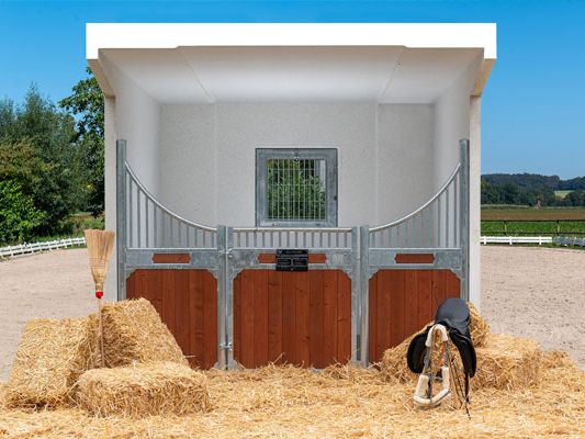 Lichte paardenstal met bruine staldeur, hooibalen en een bezem ervoor. Op de achtergrond een paardenweide.