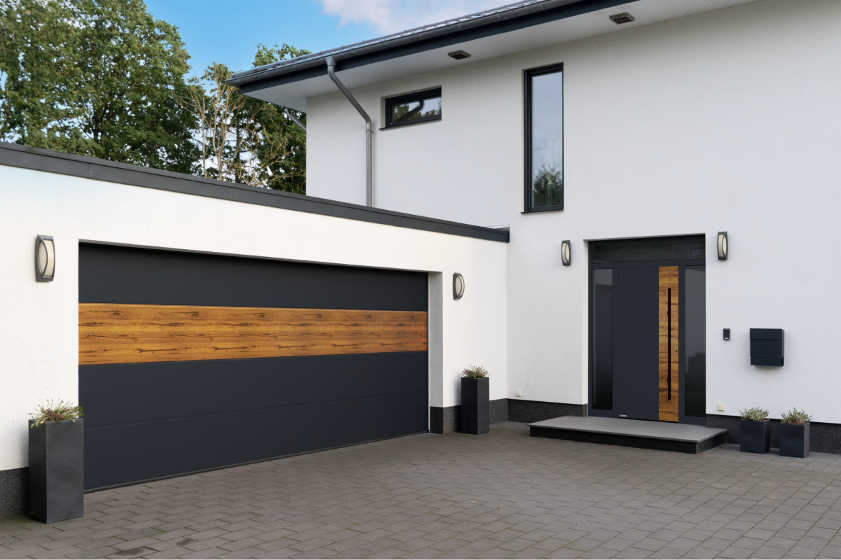 Modern huis met witte gevel, donker zadeldak en grijze garage. Voor het huis plantenbakken en bestrate binnenplaats.