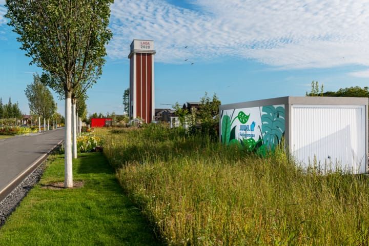 Straatbeeld met gazon, hoog gras, bomen en een hoge toren op de achtergrond. Rechts een witte gevel met plantmotieven.